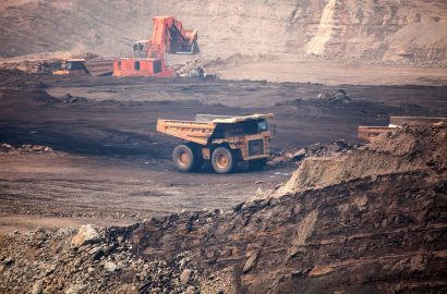 Open-cut mining site with haul truck transporting material and excavator operating in harsh, dust-heavy conditions.