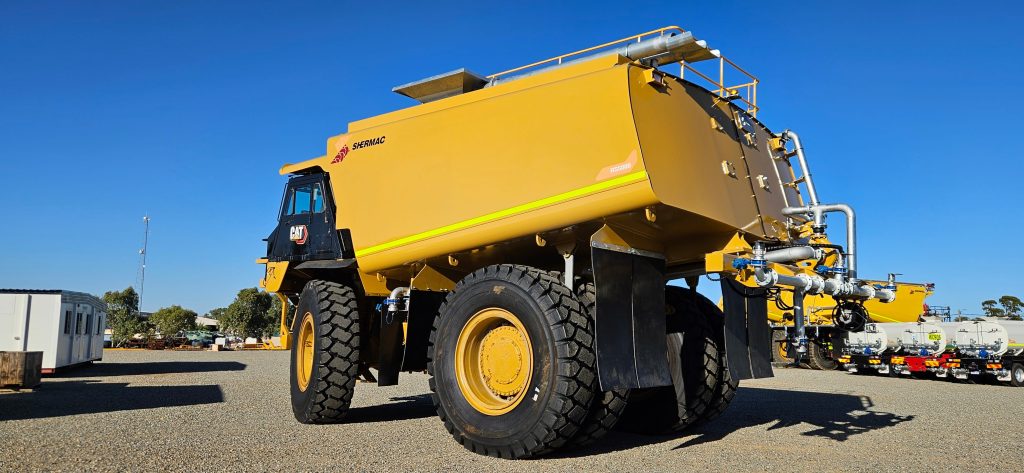 Built on a heavy-duty CAT chassis, this yellow Shermac off road water cart sits in a yard.
